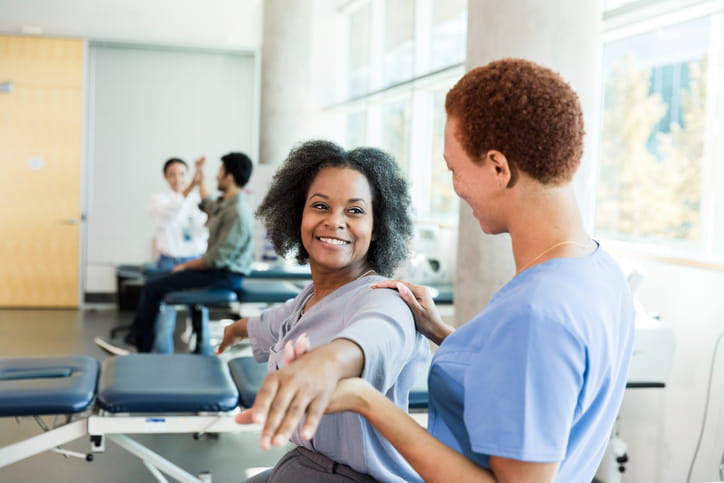 A female physical therapist in scrubs helps a smiling, middle-aged Black woman with an arm and shoulder stretching exercise in a bright rehabilitation clinic.
