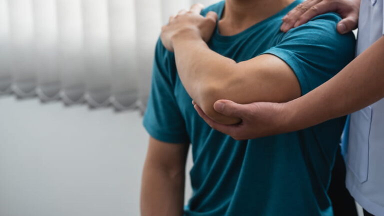 A close-up of a physical therapist's hands in a white uniform, examining a male patient's shoulder and elbow joint for pain or injury.