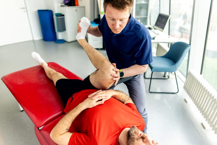 A male physical therapist performs a hip and knee stretch on a male patient who is lying on a red examination table in a clinic.