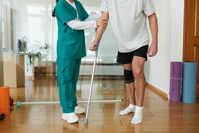 A physical therapist helps a patient who has a knee brace on and uses a crutch for assistance.